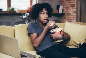 Exciting TV show. Young African man watching TV and keeping his mouth open while sitting with bucket of popcorn on the couch at home